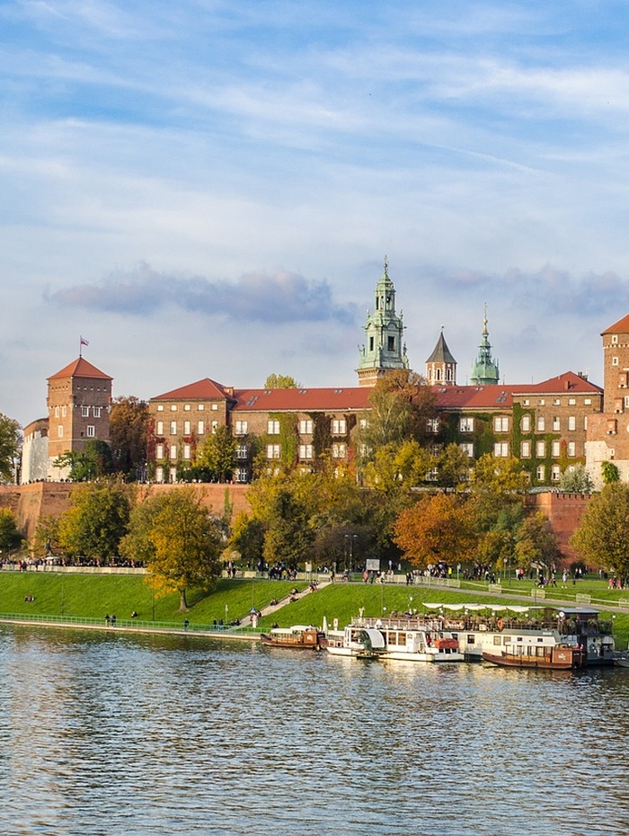 Schloss Wawel in Krakau, Polen, mit Booten auf der Weichsel und Menschen, die am Flussufer spazieren gehen.