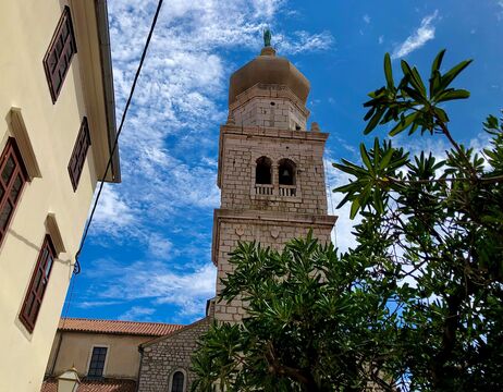 Der steinerne Kirchturm erhebt sich vor einem blauen Himmel, umrahmt von grünen Bäumen und benachbarten Gebäuden.