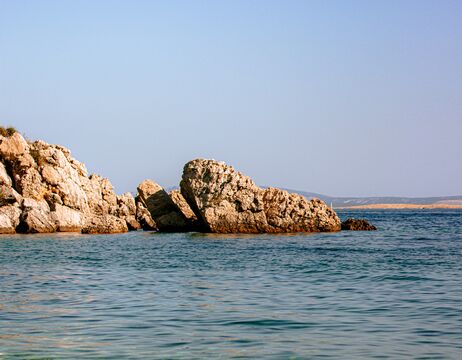 Unter einem klaren Himmel ragen große Felsen aus dem ruhigen blauen Meer, und am Horizont ist in der Ferne Land zu erkennen.
