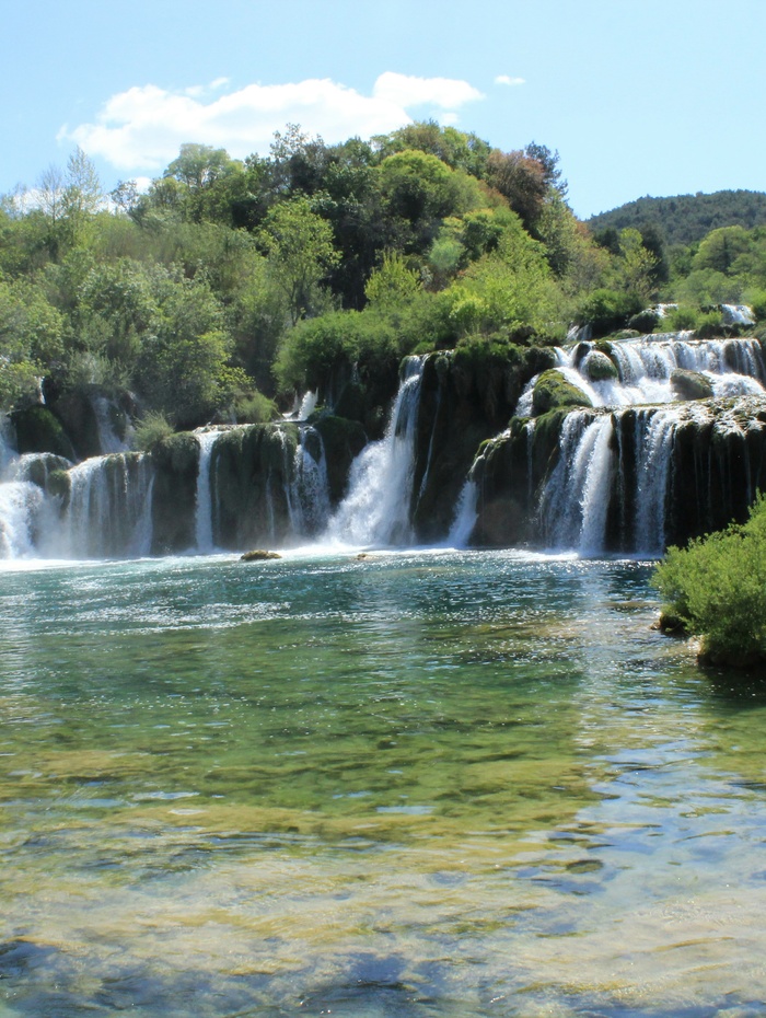 Ein Wasserfall stürzt über Felsvorsprünge in ein klares, grünlich-blaues Becken, das von üppigen Bäumen umgeben ist.
