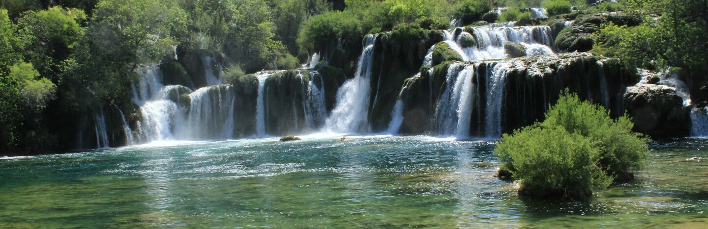 Ein Wasserfall stürzt über Felsvorsprünge in ein klares, grünlich-blaues Becken, das von üppigen Bäumen umgeben ist.