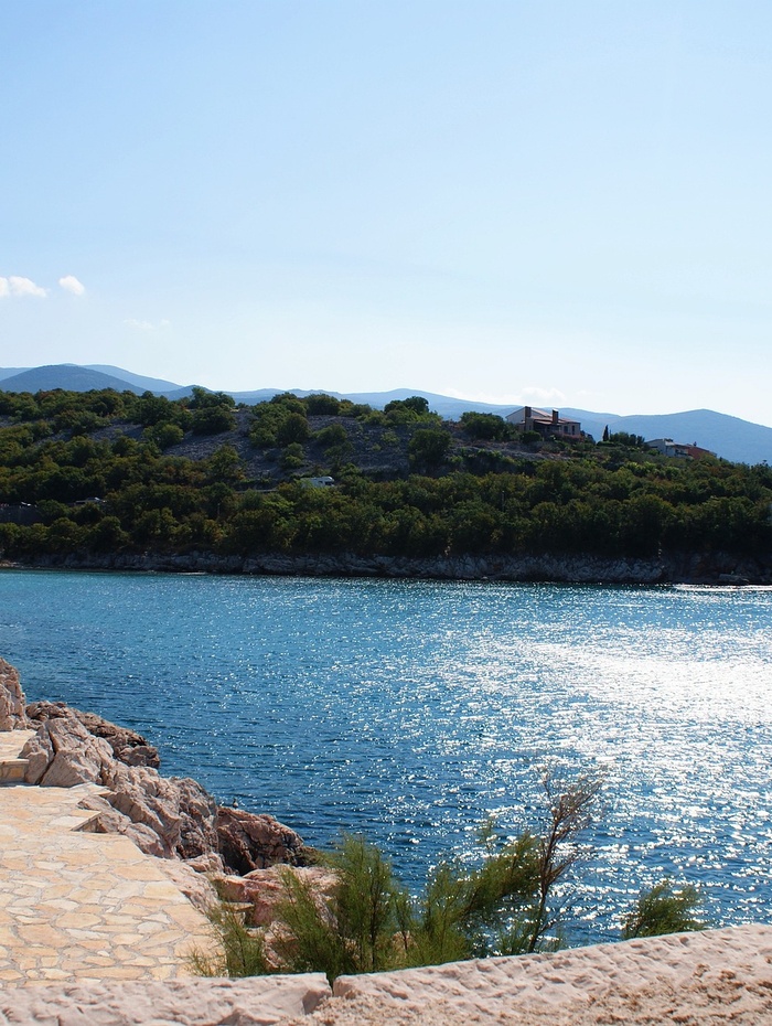 Steinweg neben einem glitzernden blauen Meer mit einem bewaldeten Hügel im Hintergrund unter einem klaren Himmel.