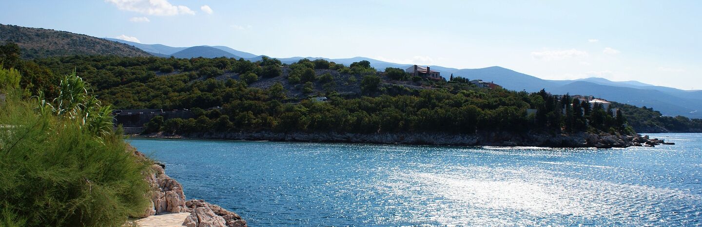 Steinweg neben einem glitzernden blauen Meer mit einem bewaldeten Hügel im Hintergrund unter einem klaren Himmel.