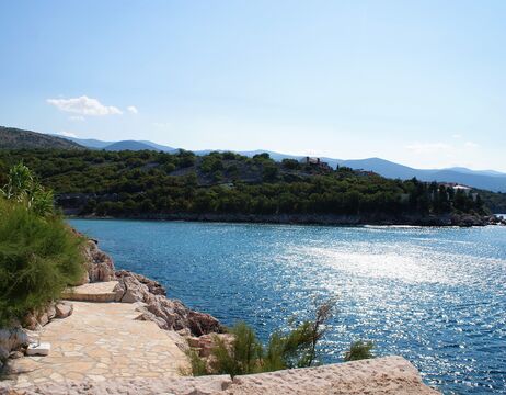 Steinweg neben einem glitzernden blauen Meer mit einem bewaldeten Hügel im Hintergrund unter einem klaren Himmel.