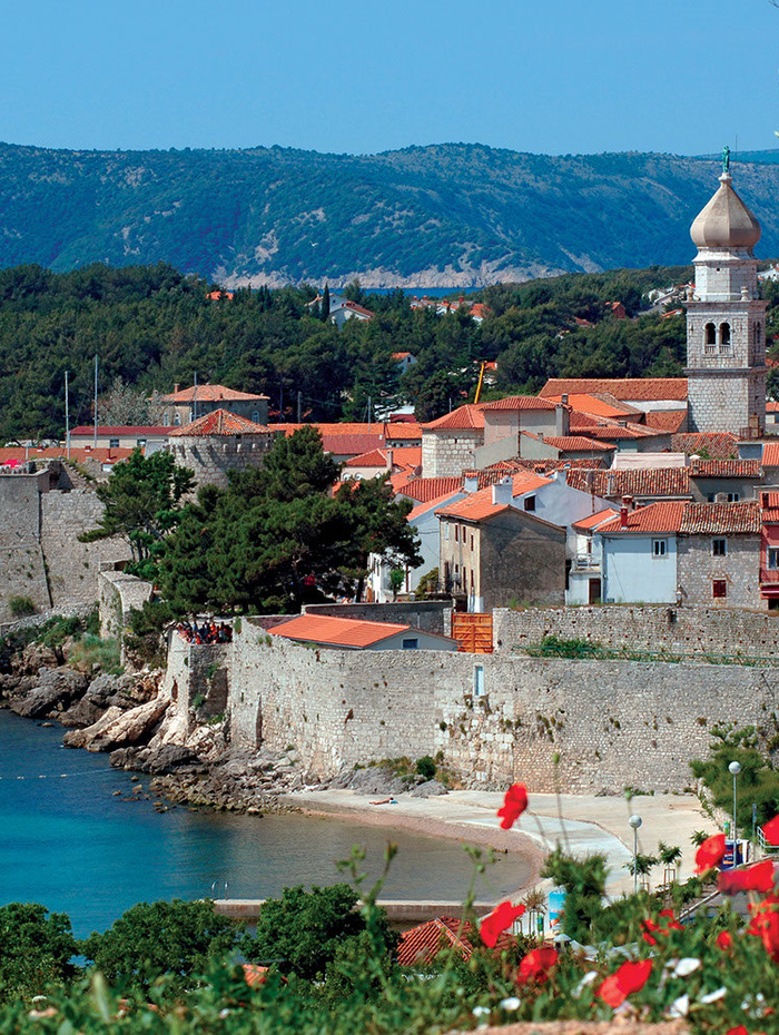 Küstenstadt mit Steinmauern, roten Ziegeldächern, einem Kirchturm und Bergen im Hintergrund.