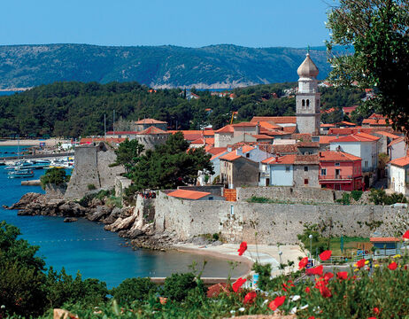 Küstenstadt mit Steinmauern, roten Ziegeldächern, einem Kirchturm und Bergen im Hintergrund.