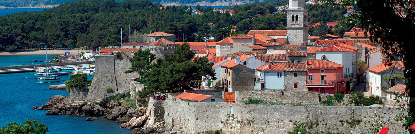Küstenstadt mit Steinmauern, roten Ziegeldächern, einem Kirchturm und Bergen im Hintergrund.