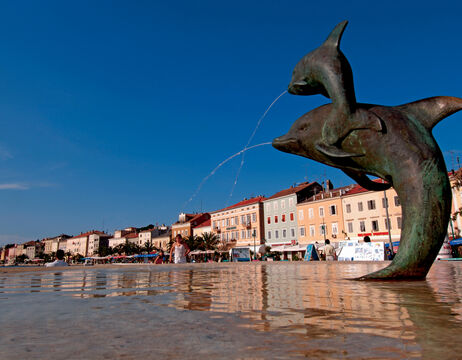 Ein bronzener Delphinbrunnen versprüht Wasser auf einem Stadtplatz am Meer mit bunten Gebäuden im Hintergrund.