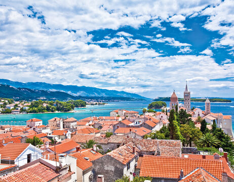Küstenstadt mit roten Dächern, Kirchtürmen, blauem Meer und Bergen unter einem teilweise bewölkten Himmel.