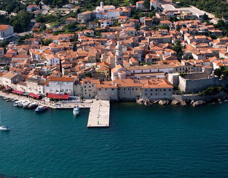 Luftaufnahme einer europäischen Küstenstadt mit roten Ziegeldächern und einem Jachthafen am blauen Meer.