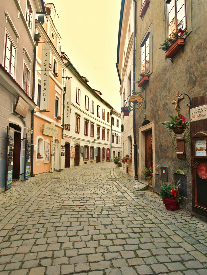 Enge Kopfsteinpflasterstraße, gesäumt von alten Gebäuden, Blumenkästen und Restaurantschildern an einem ruhigen Tag.