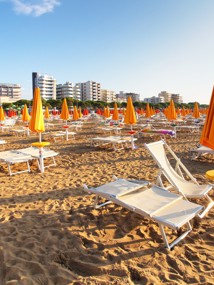 Sandstrand mit Reihen leerer Liegestühle und orangefarbener Sonnenschirme, Gebäude im Hintergrund.