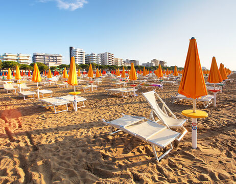 Sandstrand mit Reihen leerer Liegestühle und orangefarbener Sonnenschirme, Gebäude im Hintergrund.