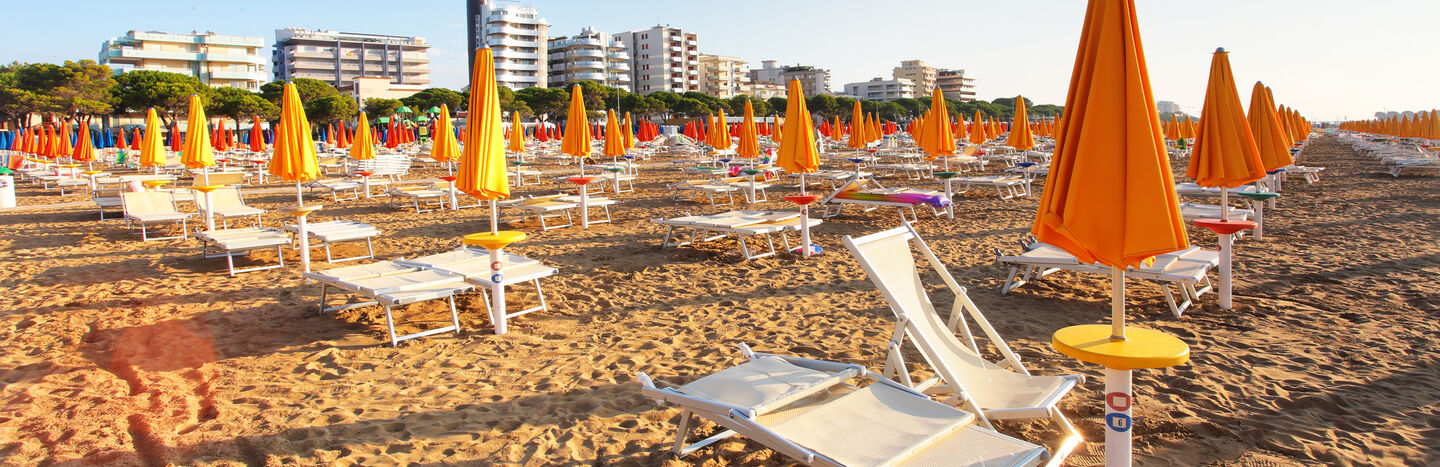 Sandstrand mit Reihen leerer Liegestühle und orangefarbener Sonnenschirme, Gebäude im Hintergrund.