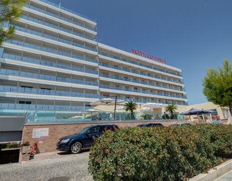 Das mehrstöckige Hotel Caesarus mit Balkonen, Pool, geparkten Autos und Grünanlagen unter einem strahlend blauen Himmel.