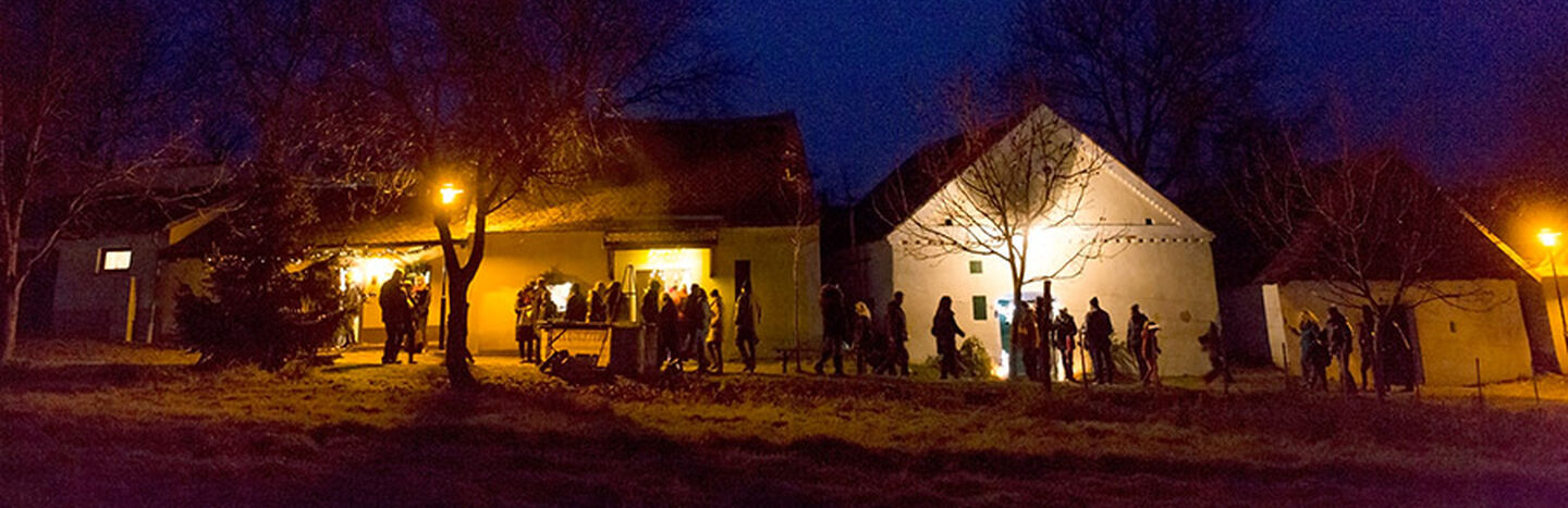 Die Menschen versammeln sich nachts vor warm beleuchteten Häusern unter einem dunkelblauen Himmel, in dessen Nähe kahle Bäume stehen.