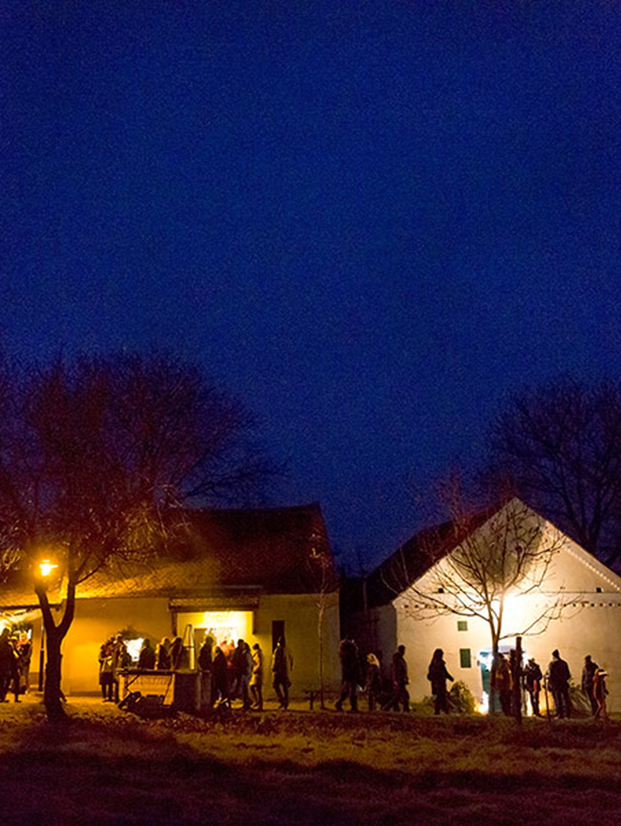 Die Menschen versammeln sich nachts vor warm beleuchteten Häusern unter einem dunkelblauen Himmel, in dessen Nähe kahle Bäume stehen.