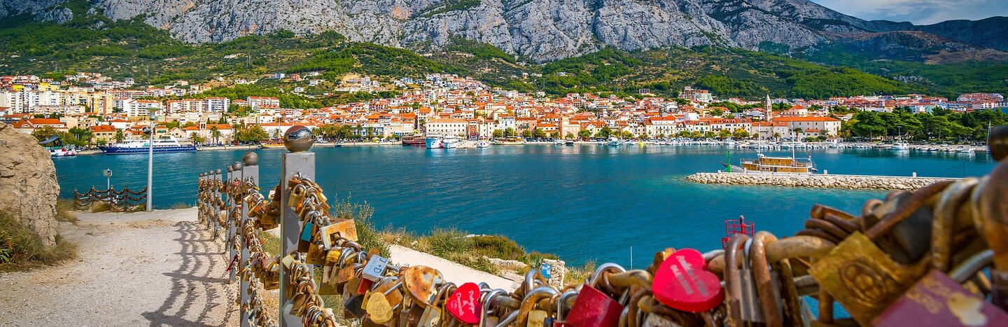 Bunte Vorhängeschlösser an einem Zaun überblicken eine Stadt am Meer mit Bergen im Hintergrund unter einem teilweise bewölkten Himmel.