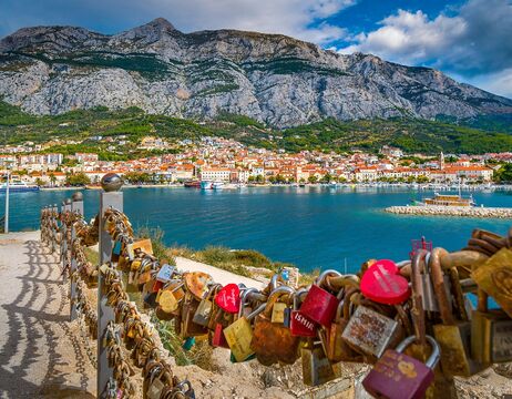 Bunte Vorhängeschlösser an einem Zaun überblicken eine Stadt am Meer mit Bergen im Hintergrund unter einem teilweise bewölkten Himmel.