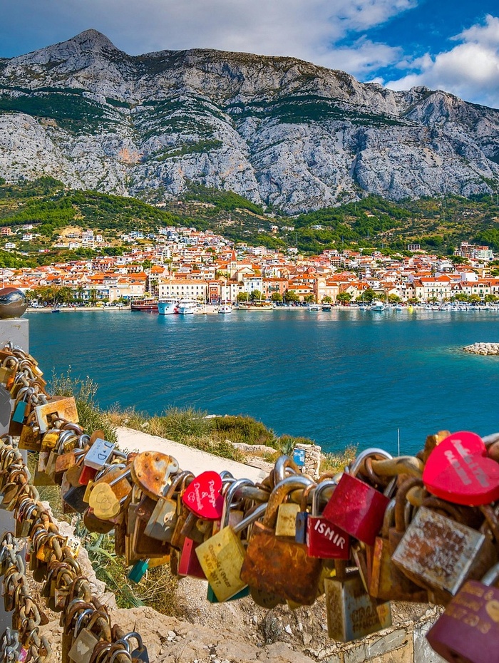 Bunte Vorhängeschlösser an einem Zaun überblicken eine Stadt am Meer mit Bergen im Hintergrund unter einem teilweise bewölkten Himmel.