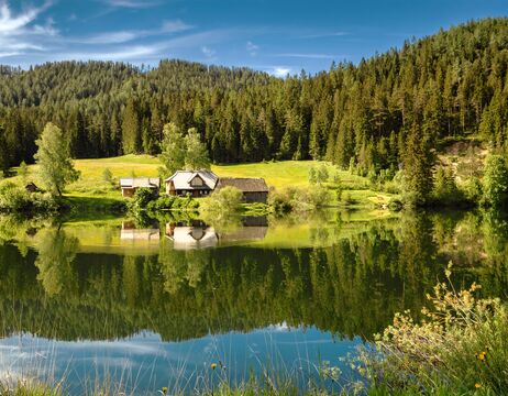 Kleine Häuser an einem See mit Wald und Bergen im Hintergrund, die sich im ruhigen Wasser spiegeln.