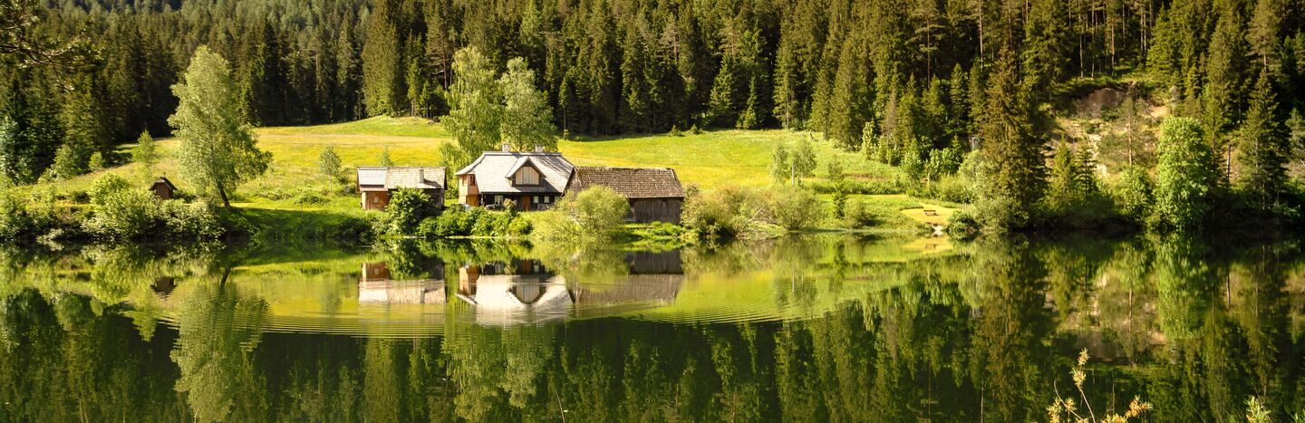 Kleine Häuser an einem See mit Wald und Bergen im Hintergrund, die sich im ruhigen Wasser spiegeln.