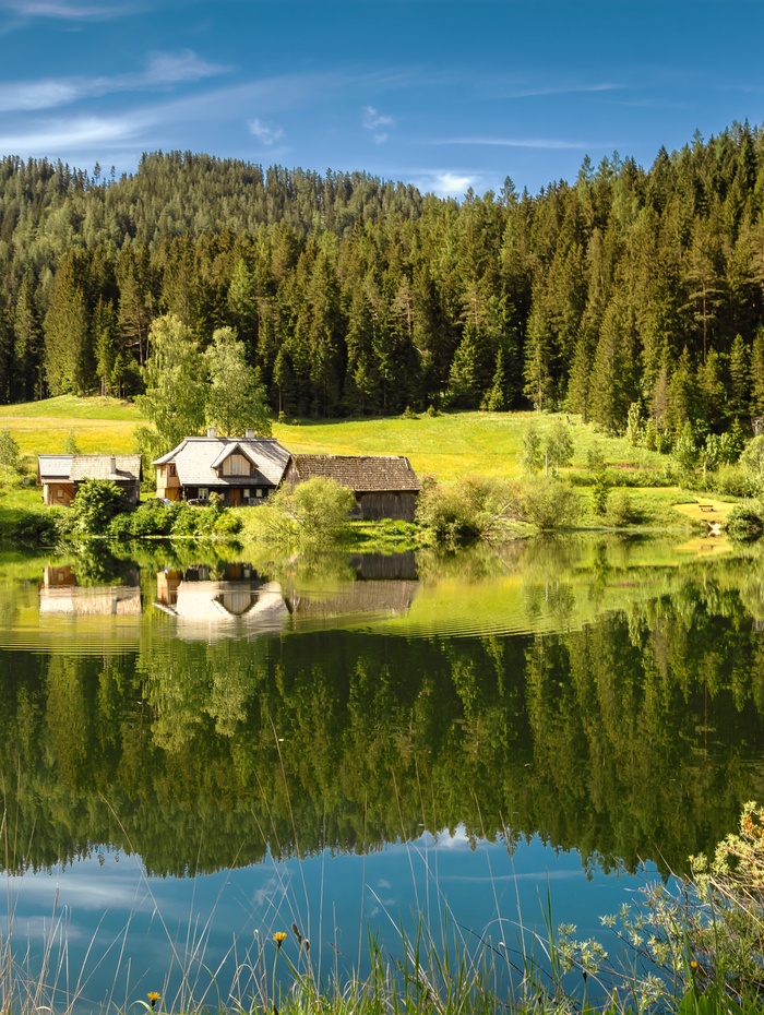 Kleine Häuser an einem See mit Wald und Bergen im Hintergrund, die sich im ruhigen Wasser spiegeln.