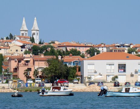 Kleine Boote auf ruhigem Wasser mit orangefarbenen Dächern und einem hohen weißen Kirchturm im Hintergrund.