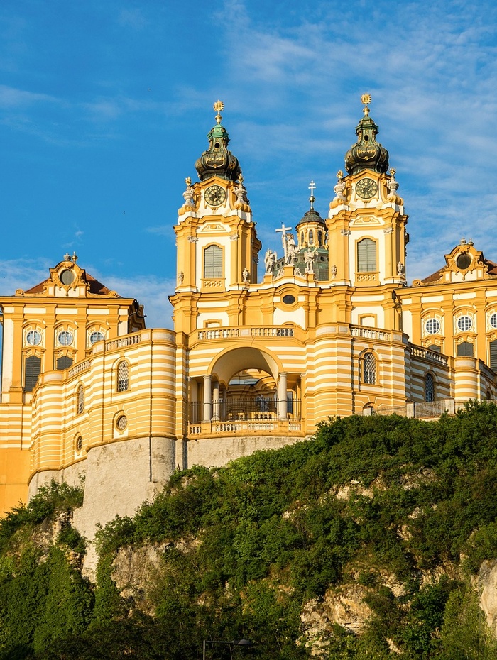 Gelbe Barockabtei mit Zwillingstürmen auf einem Hügel vor einem blauen Himmel mit einigen Wolken.