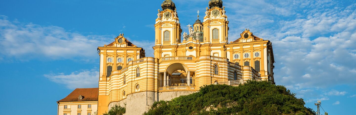 Gelbe Barockabtei mit Zwillingstürmen auf einem Hügel vor einem blauen Himmel mit einigen Wolken.