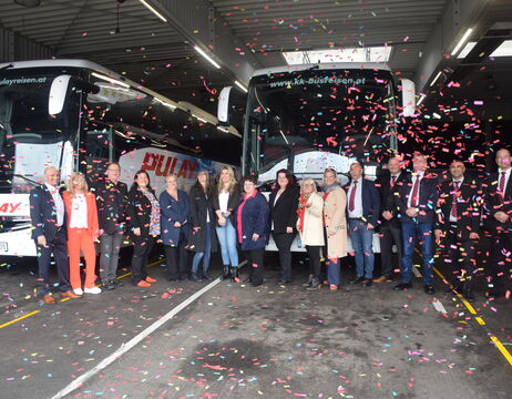 Eine Gruppe von Menschen steht lächelnd zwischen Bussen in einer Garage, umgeben von fallendem Konfetti.
