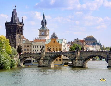 Historische Steinbrücke mit Türmen über einen Fluss, mit bunten Gebäuden und blauem Himmel im Hintergrund.