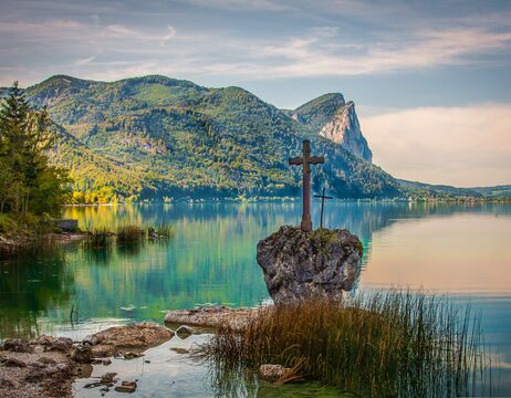 Ein Steinkreuz steht an einem ruhigen See mit grünen Bergen im Hintergrund unter einem blauen Himmel.