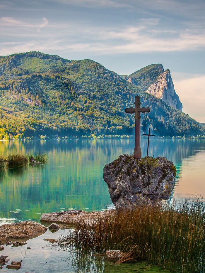 Ein Steinkreuz steht an einem ruhigen See mit grünen Bergen im Hintergrund unter einem blauen Himmel.