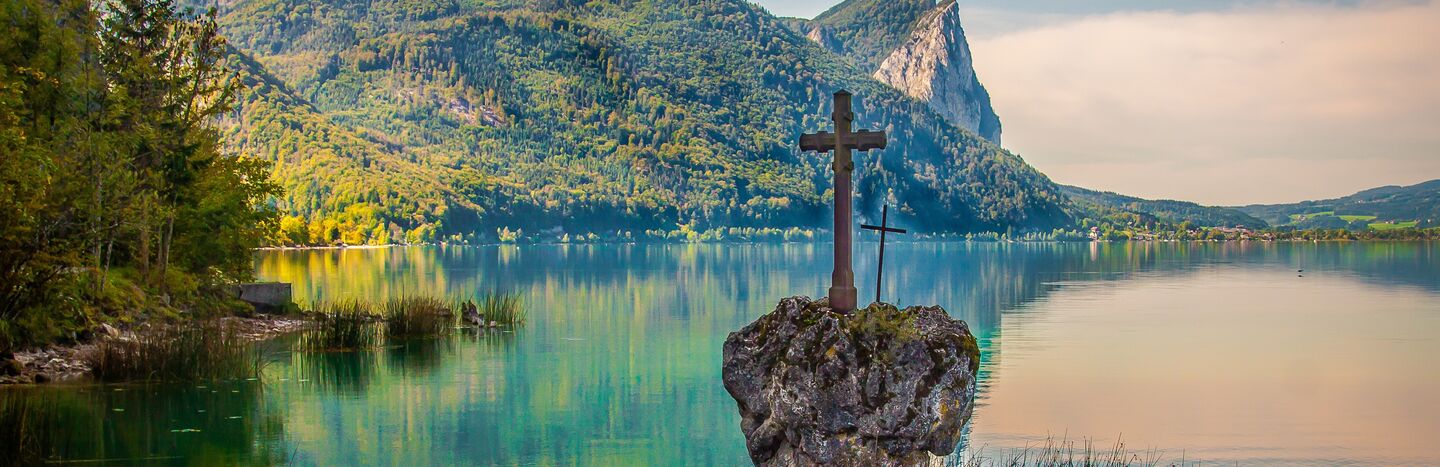 Ein Steinkreuz steht an einem ruhigen See mit grünen Bergen im Hintergrund unter einem blauen Himmel.