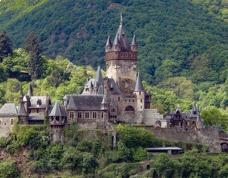 Eine mittelalterliche Burg mit hohen Türmen thront inmitten üppiger grüner Bäume am Fuße eines bewaldeten Hügels.