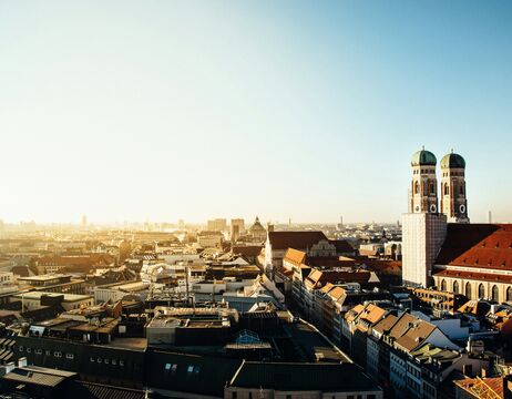 Ein Stadtbild bei Sonnenaufgang mit historischen Gebäuden und roten Dächern unter einem klaren blauen Himmel.