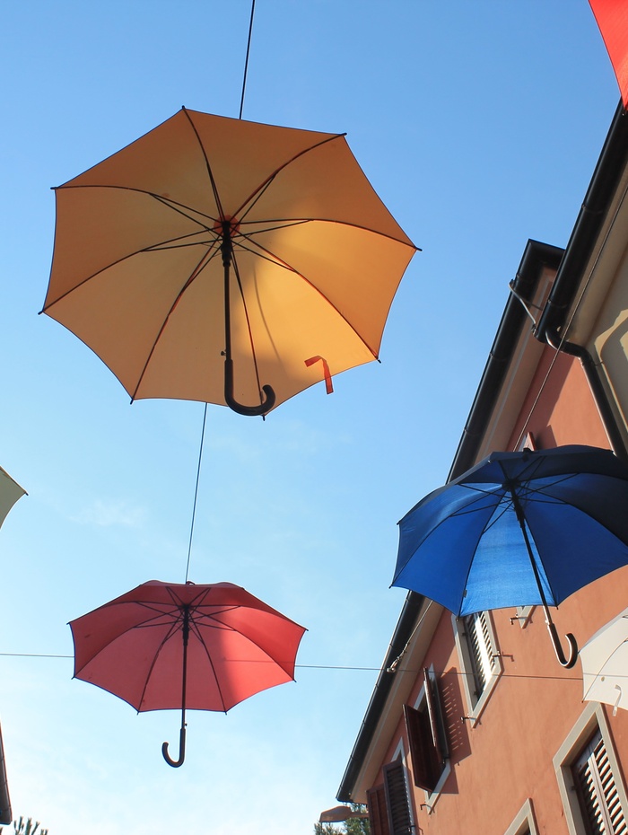 Drei bunte Regenschirme hängen über einer Straße zwischen Gebäuden unter einem klaren blauen Himmel.