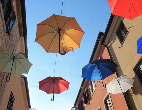 Drei bunte Regenschirme hängen über einer Straße zwischen Gebäuden unter einem klaren blauen Himmel.