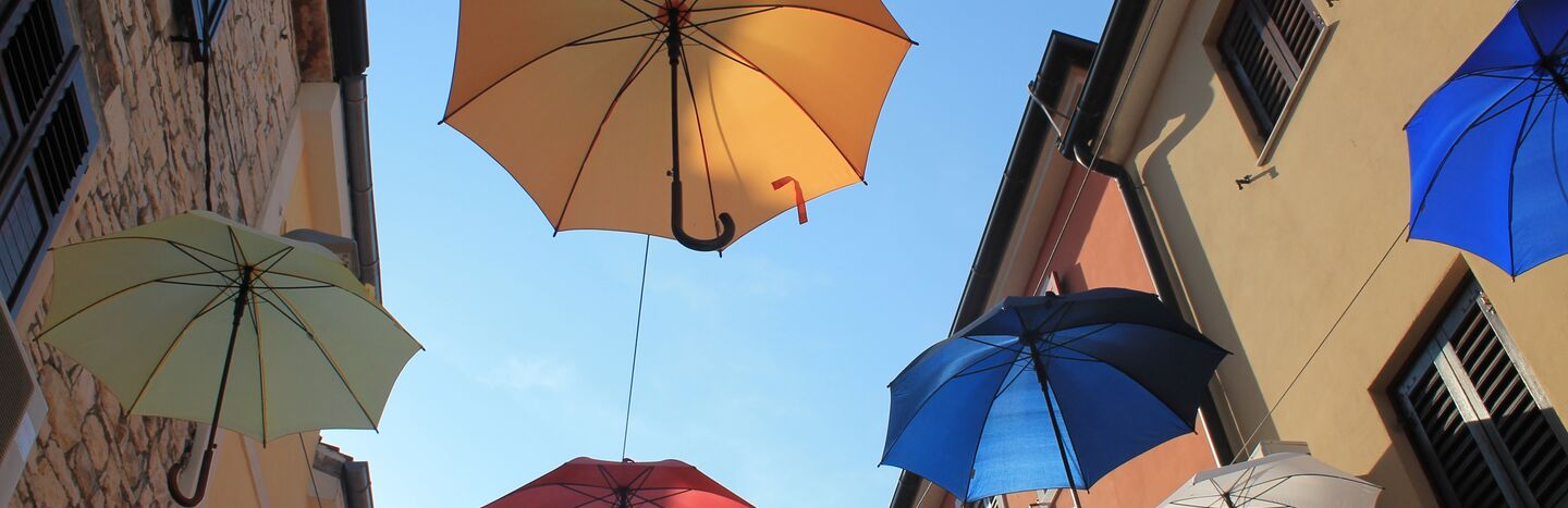 Drei bunte Regenschirme hängen über einer Straße zwischen Gebäuden unter einem klaren blauen Himmel.