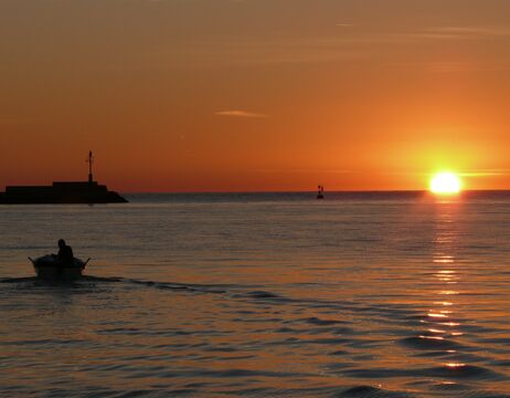 Eine Person rudert ein Boot auf ruhigem Wasser bei Sonnenuntergang, die Sonne steht nahe am Horizont und der Himmel ist orange.