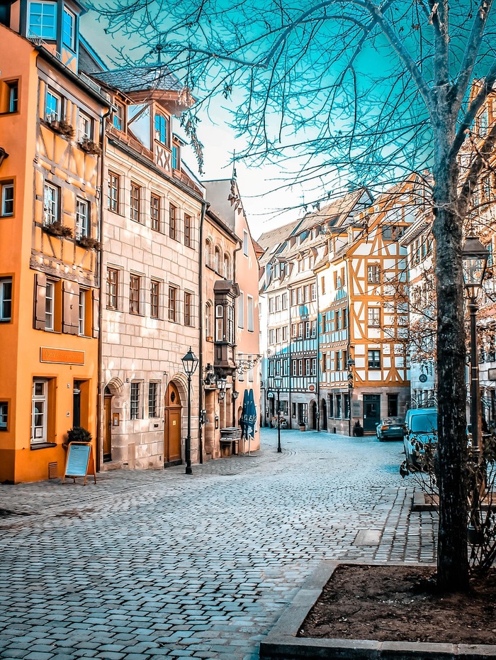 Eine Straße mit Kopfsteinpflaster, gesäumt von bunten, alten europäischen Gebäuden und kahlen Bäumen unter einem strahlend blauen Himmel.