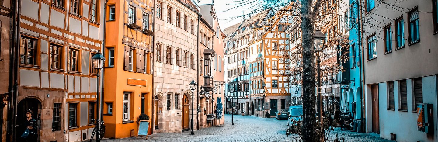 Eine Straße mit Kopfsteinpflaster, gesäumt von bunten, alten europäischen Gebäuden und kahlen Bäumen unter einem strahlend blauen Himmel.