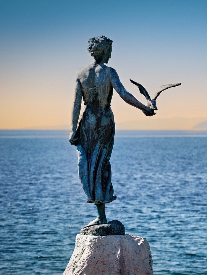 Luftaufnahme einer Küstenstadt mit roten Dächern, einem Glockenturm, einem Jachthafen und blauem Wasser.