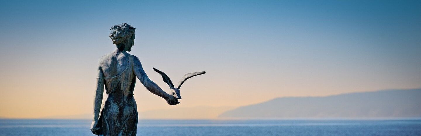 Luftaufnahme einer Küstenstadt mit roten Dächern, einem Glockenturm, einem Jachthafen und blauem Wasser.