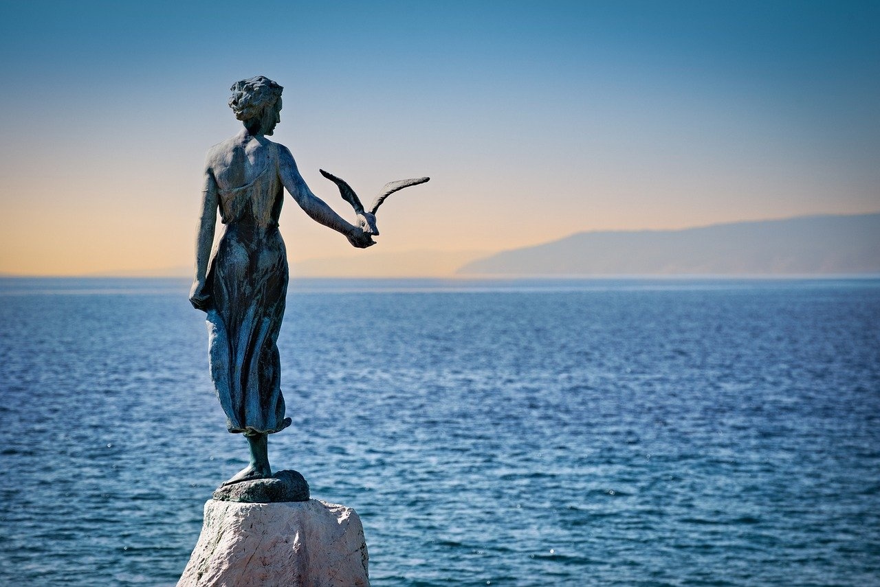 Eine Statue am Meer mit einer Frau, die einen Vogel hält, mit Blick auf ruhiges blaues Wasser und ferne Berge.
