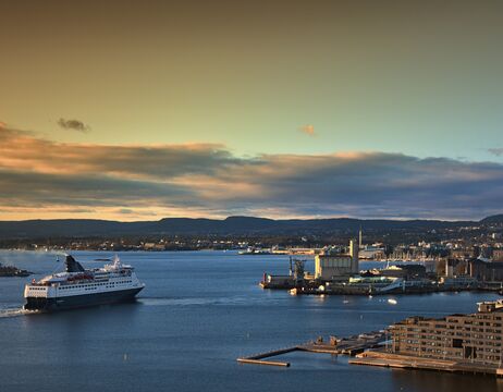 Ein Kreuzfahrtschiff nähert sich einem Hafen bei Sonnenuntergang, mit entfernten Hügeln und bewölktem Himmel im Hintergrund.