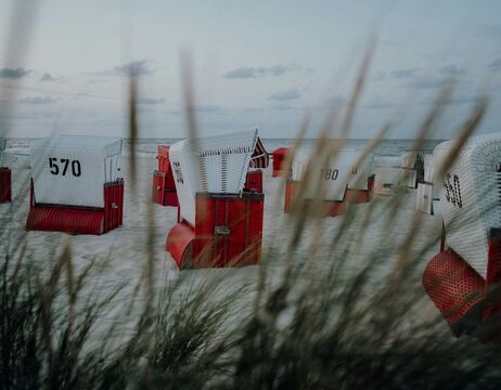 Rot-weiße Strandkörbe mit Nummern stehen auf dem Sand, durch hohes Strandgras in der Nähe des Wassers betrachtet.