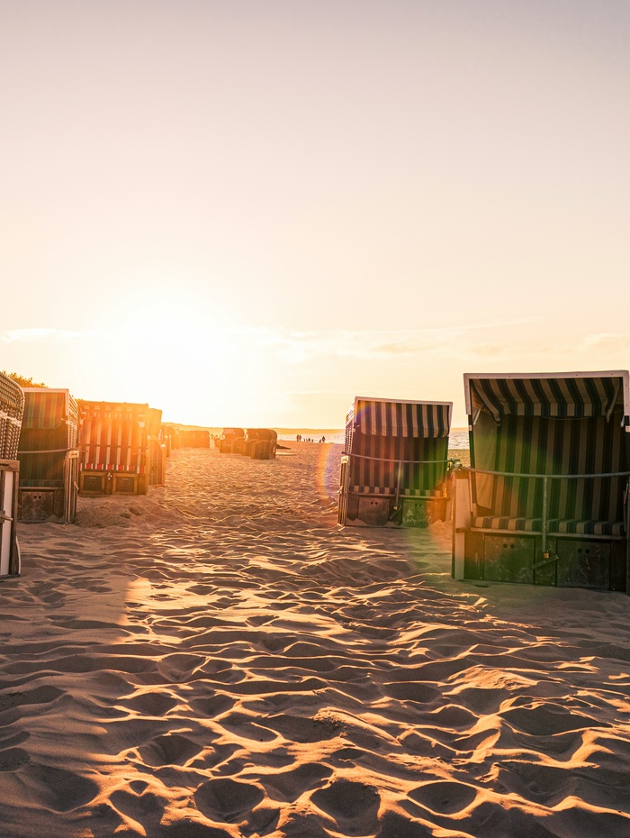 Sonnenuntergang über dem Sandstrand mit gestreiften Strandkörben, die den Weg zum Meer säumen.