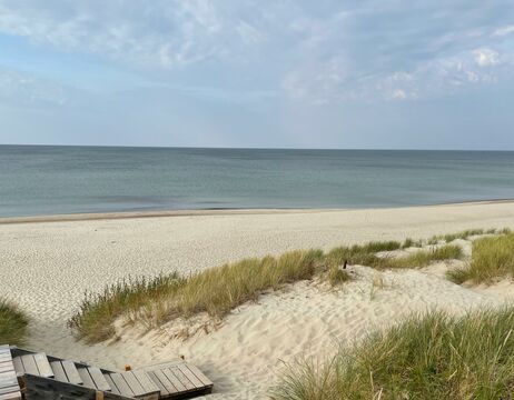 Holzstufen führen zu einem Sandstrand mit grasbewachsenen Dünen und ruhigem, blauem Wasser unter einem teilweise bewölkten Himmel.